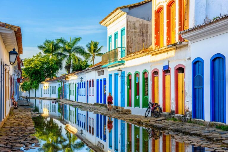 A imagem mostra uma rua pitoresca de Paraty, no estado do Rio de Janeiro, Brasil. As fachadas coloniais dos edifícios são pintadas em tons vibrantes, como azul, amarelo, vermelho e verde, criando uma atmosfera charmosa e colorida. O chão é formado por pedras irregulares, características da arquitetura colonial, e a rua está levemente alagada, refletindo as construções e o céu. Há algumas pessoas caminhando e uma bicicleta encostada, reforçando a tranquilidade do cenário. Ao fundo, palmeiras complementam o ambiente tropical. A iluminação natural destaca a beleza das cores e o aspecto histórico do lugar.